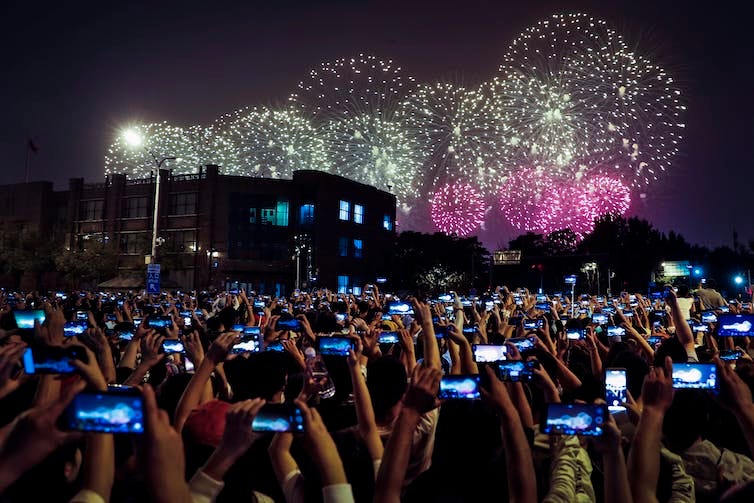 Thousands of people at a night-time celebration hold up their smartphones to take pictures of fireworks.