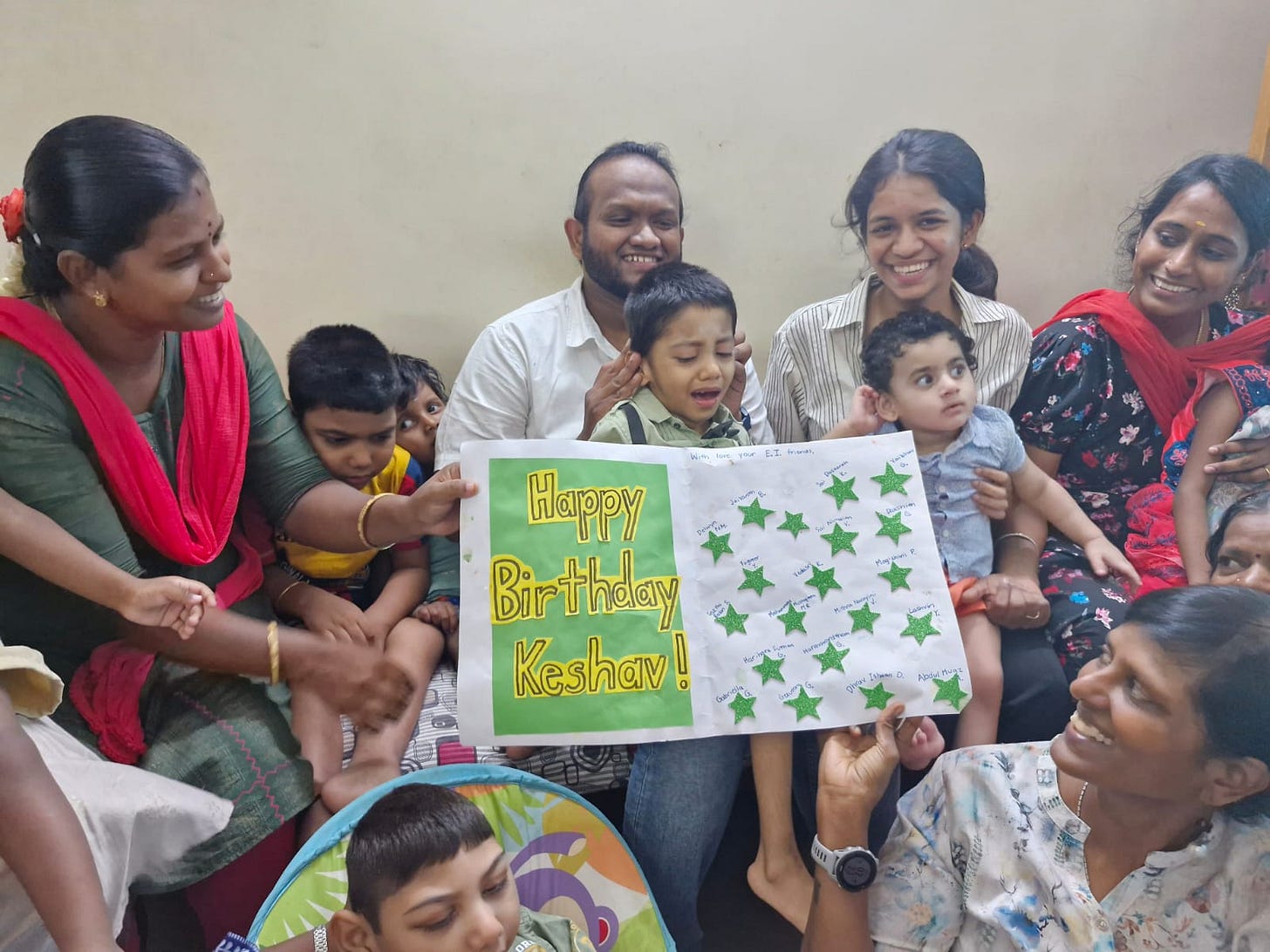 The image captures a joyful celebration of Keshav's 5th birthday. Keshav, seated in the center, is surrounded by six of his classmates, their mothers, and grandmothers, and his teachers from Vidya Sagar. Everyone is smiling and happy, celebrating this special occasion together. A large handmade card with the words "Happy Birthday Keshav!" decorated with green stars is prominently displayed. The atmosphere is filled with warmth and happiness as they wish Keshav well on his special day.