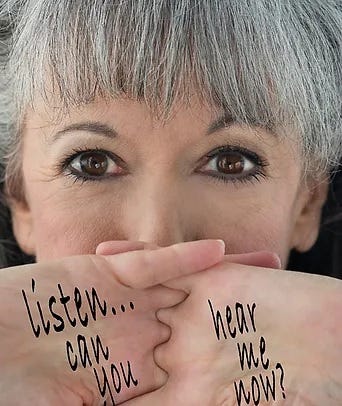 Gloria Rosen looking at the camera covering her mouth with her hands. The words "listen... can you hear me now?" are written on her hands