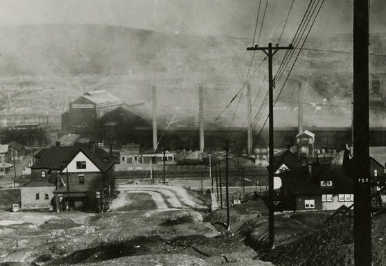 view down a hill toward a town, smokestacks, buildings, houses, lots of smoke