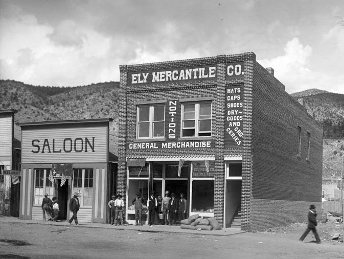 Old black and white photos of two buildings in Ely, Nevada. On the left is a one-story saloon, with the word "SALOON" painted in large letters on the top of the wooden building. To the right is a two-story brick building with the words, "ELY MERCANTILE CO." painted in white letters along the top. The are various men standing and walking in front of the two buildings. A large mountain sits in the background. The road is unpaved and a vacant lots sits to the right.