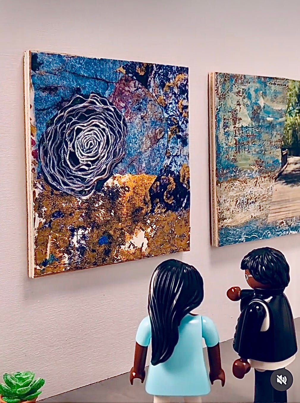 A micro gallery with white walls and two mixed media pieces. They are both colorful abstracts with photographs. One has a picture of a large rosette made from vines and the other a wooden boardwalk that looks like it curves into the mists of time.