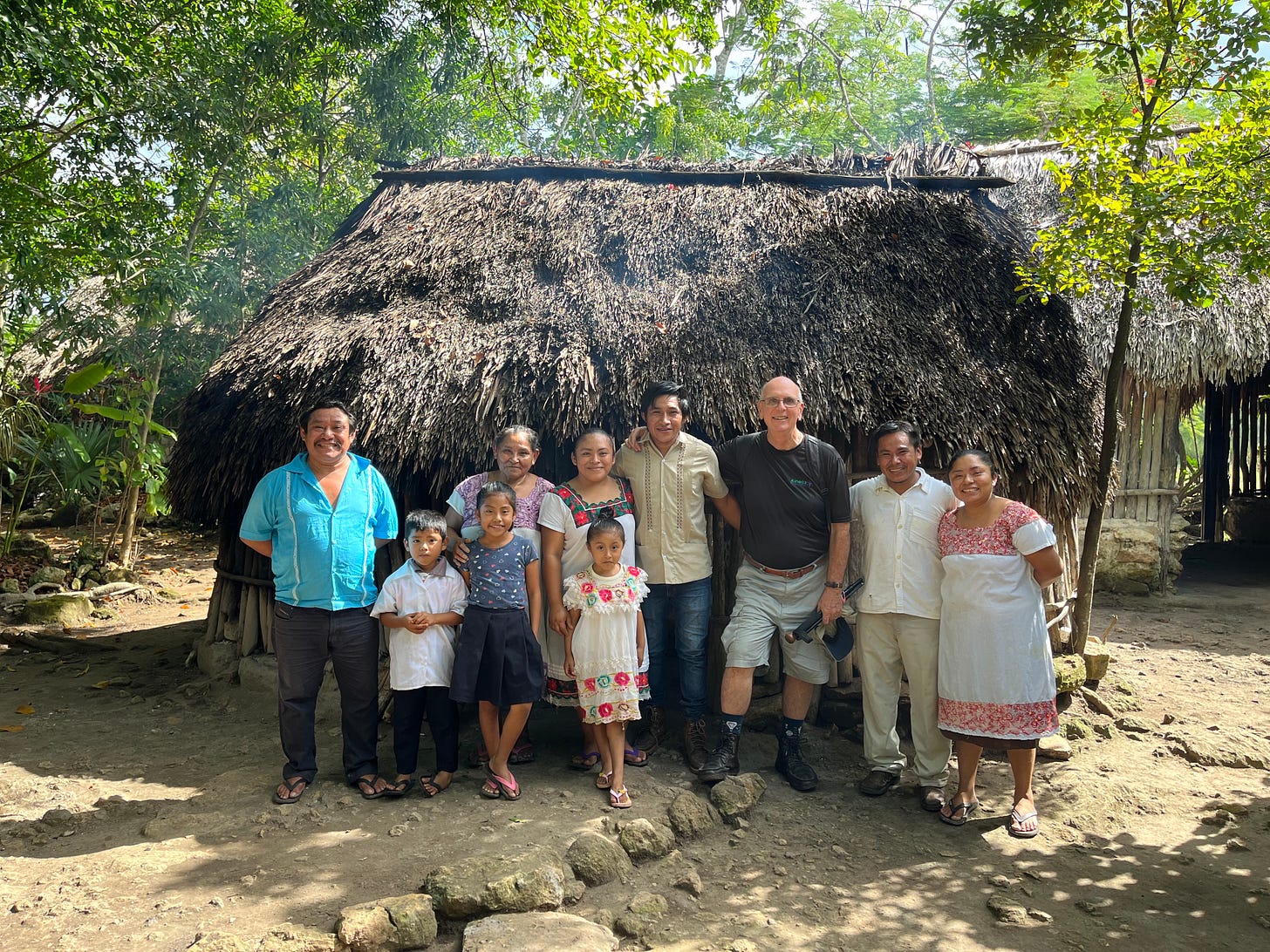 Multi-generational family, some in traditional dress, standing in front of their thatched roof home called a pilapa.