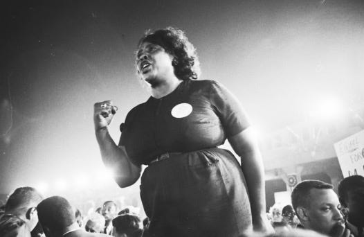 A Black disabled woman seems to float above a crowd, holding a fist of resistance close to her body, in the middle of crying out.