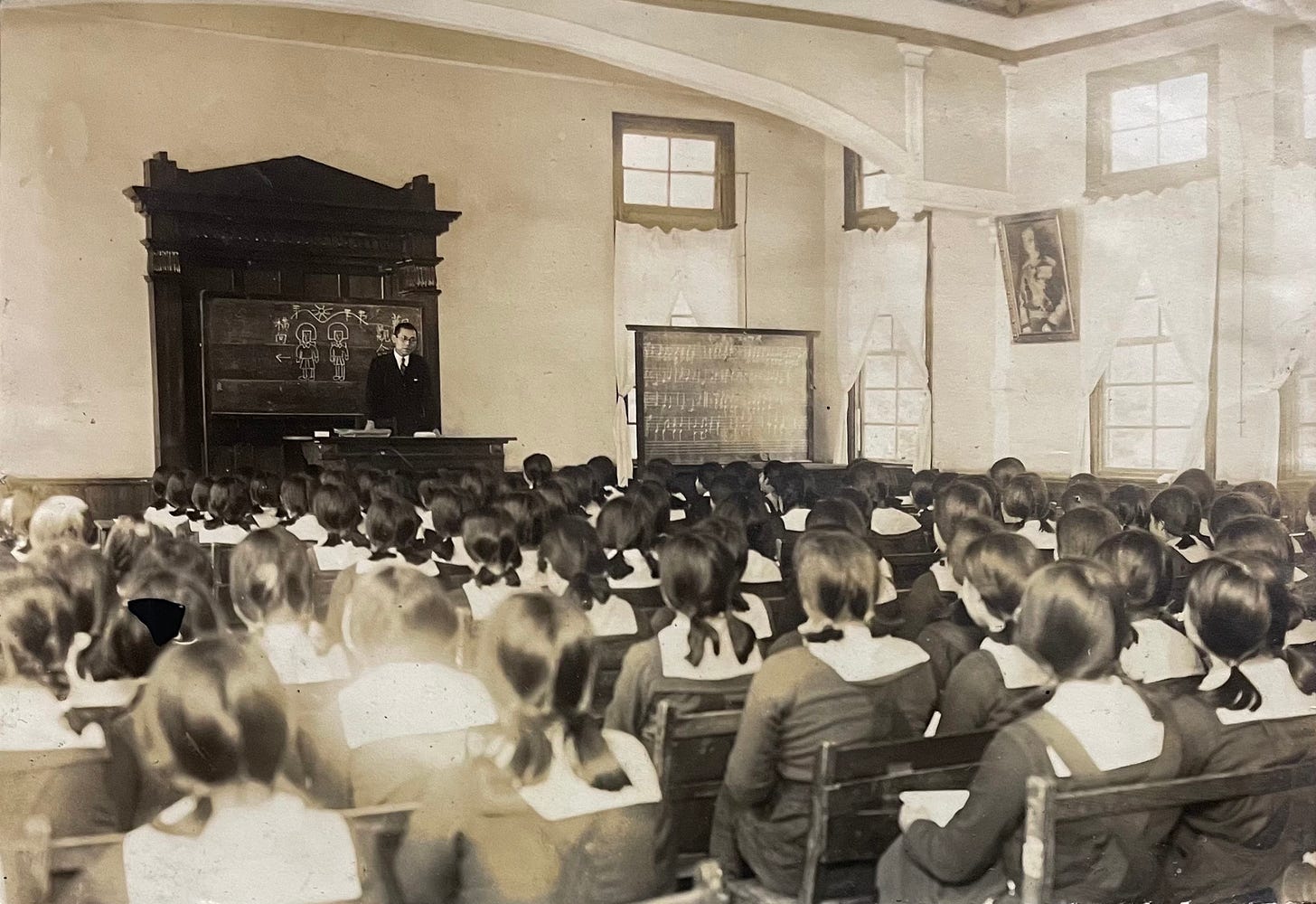 A black and white photo of a large classroom filled with young girls all in school uniforms. At the front of the classroom stands man in a suit standing in front of a drawing of two figures.