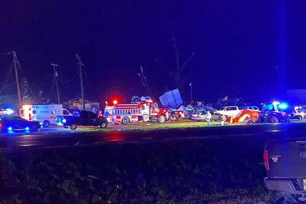 Fire trucks and ambulances along with other vehicles line a road at night, with damaged buildings and downed power lines behind them.