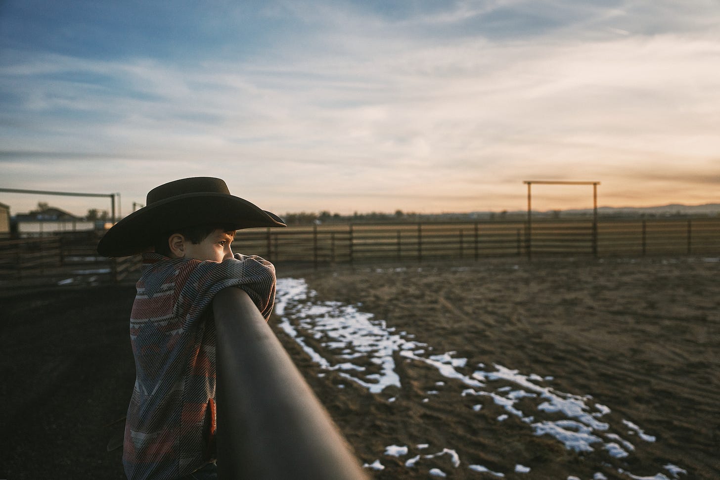 A boy in a cowboy hat overlooks a horse riding arena at sunset