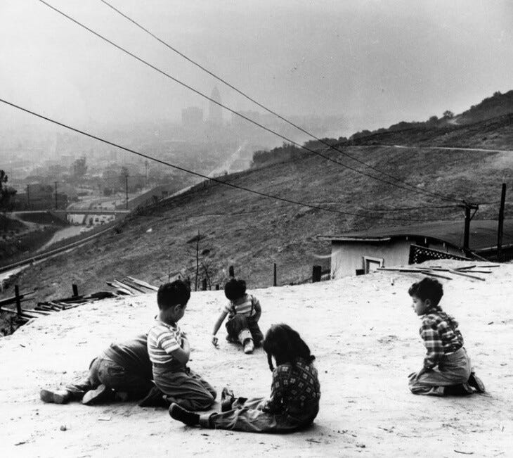 Five children sit on the ground playing. An empty hillside is in the near background and the bare outlines of a city skyline is beyond.