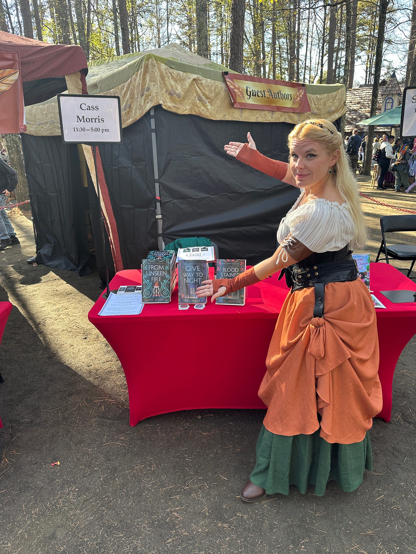 Cass, dressed in medieval-ish clothing of a fluffy blouse, leather corset, orange skirt gathered with skirt hikes, a green underskirt, and her multi-pouch hip belt, gesturing at a table stacked with her books. A tent behind the table proclaims GUEST AUTHORS, and the rest of the background is a bustling street of a Reniassance faire.