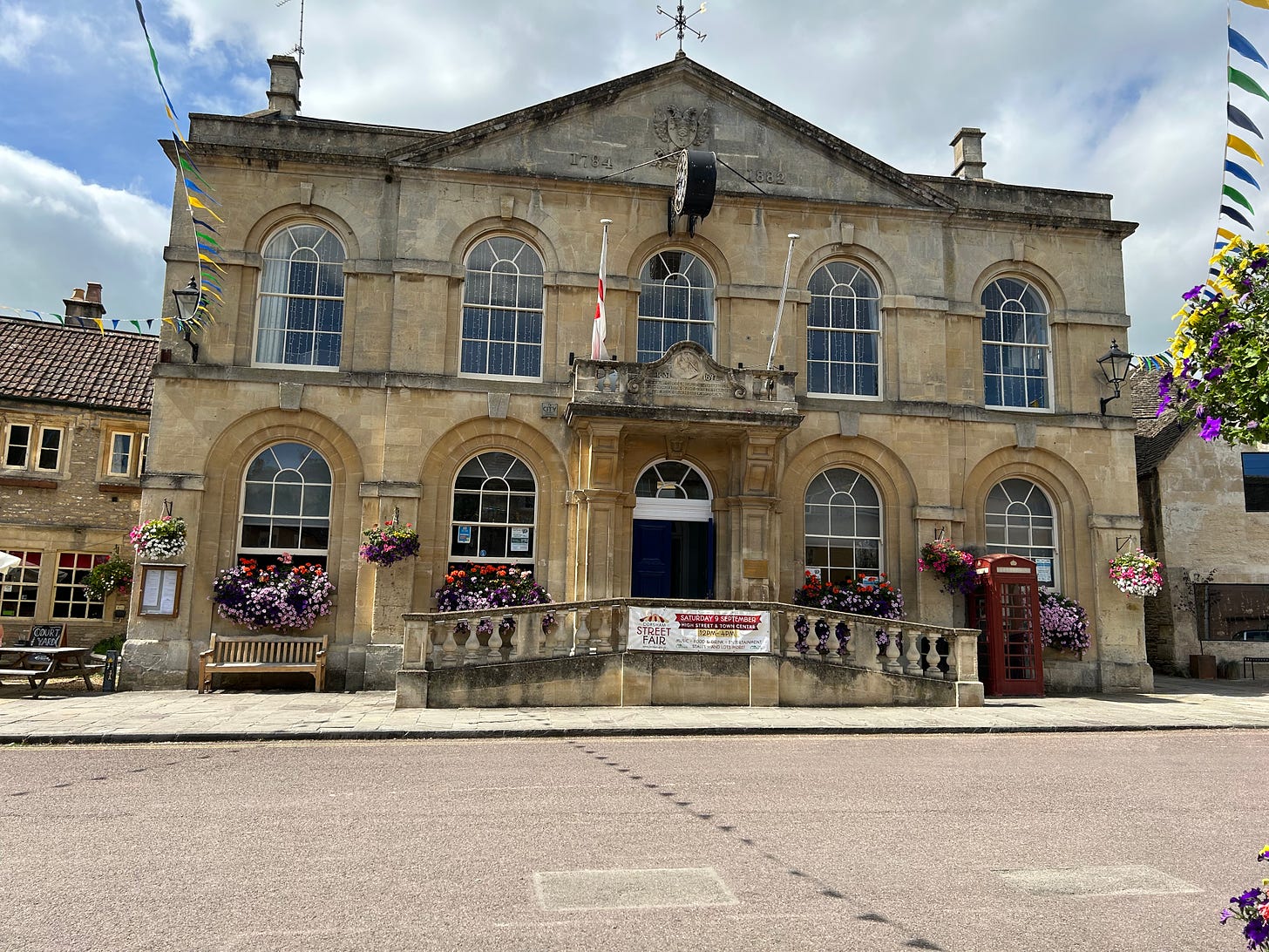 The stone built Corsham Town Hall. 4 windows on the ground floor with a central doorway and 5 windows on the first floor. A large clock is hung over the top middle window facing down the street rather than out from the building. Image: Roland's Travels