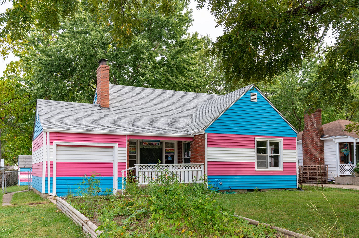 A one story house with the siding painted in stripes of white, pink, and blue, resembling the transgender pride flag.