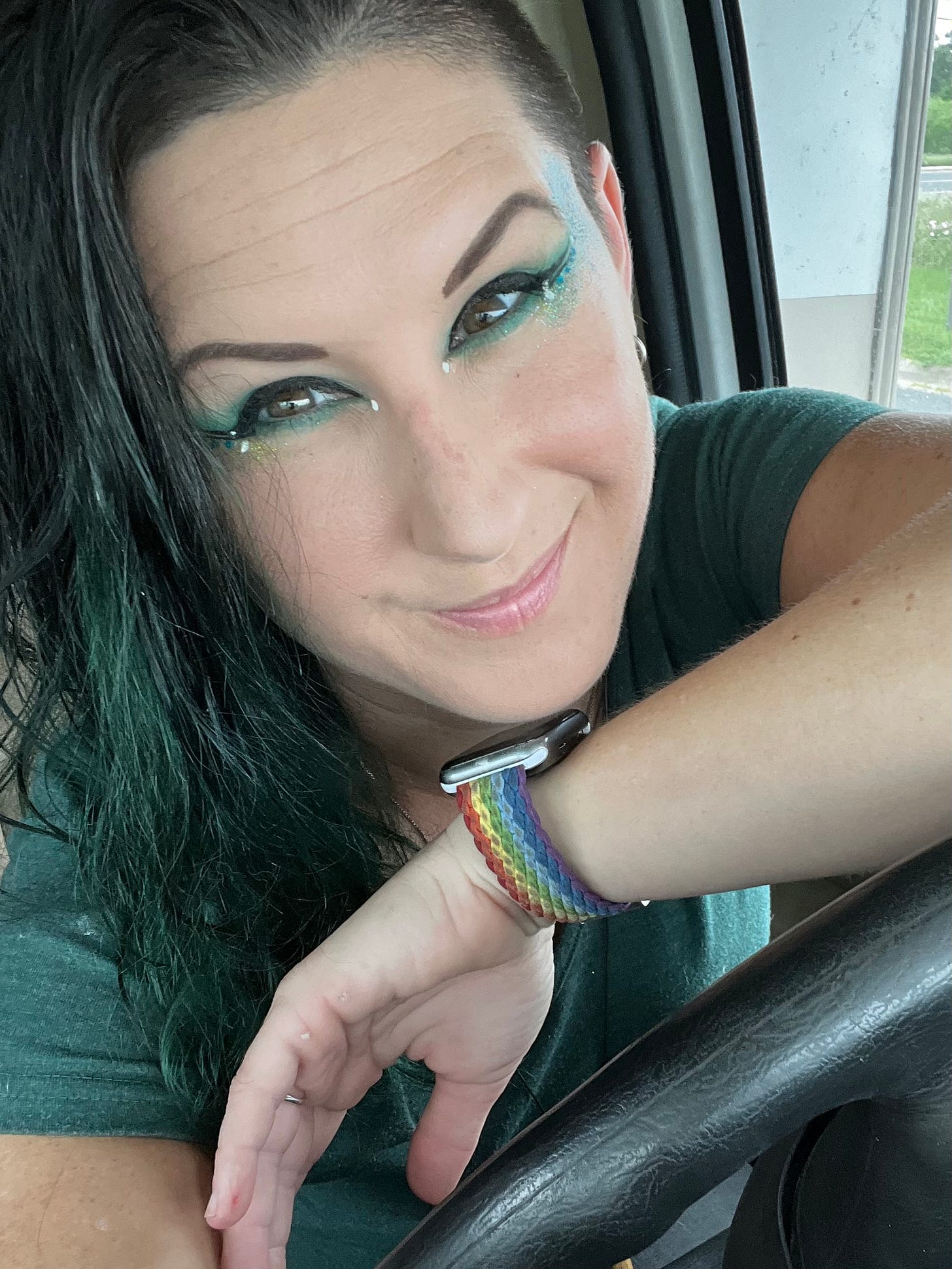This is a photo of Lyric, dressed in Green for spring. They have bold green, black, and white-winged eyeliner and a green shirt. They are leaning on the RV's steering wheel with their left arm, which has a rainbow watch on it.