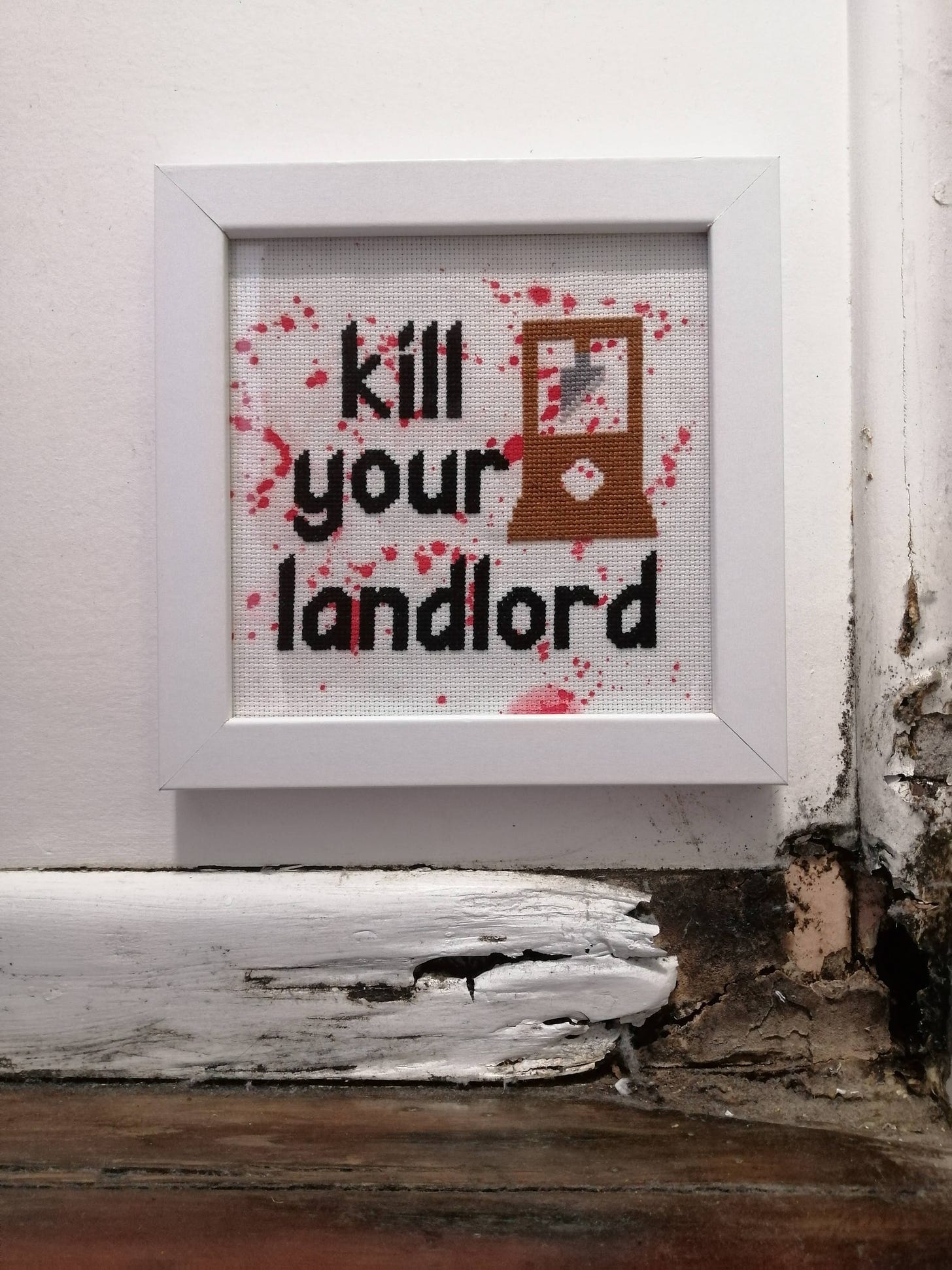 Cross stitch says "kill your landlord" on a red splattered fabric with a picture of a guillotine. The frame is in a mouldy rotting corner of a house