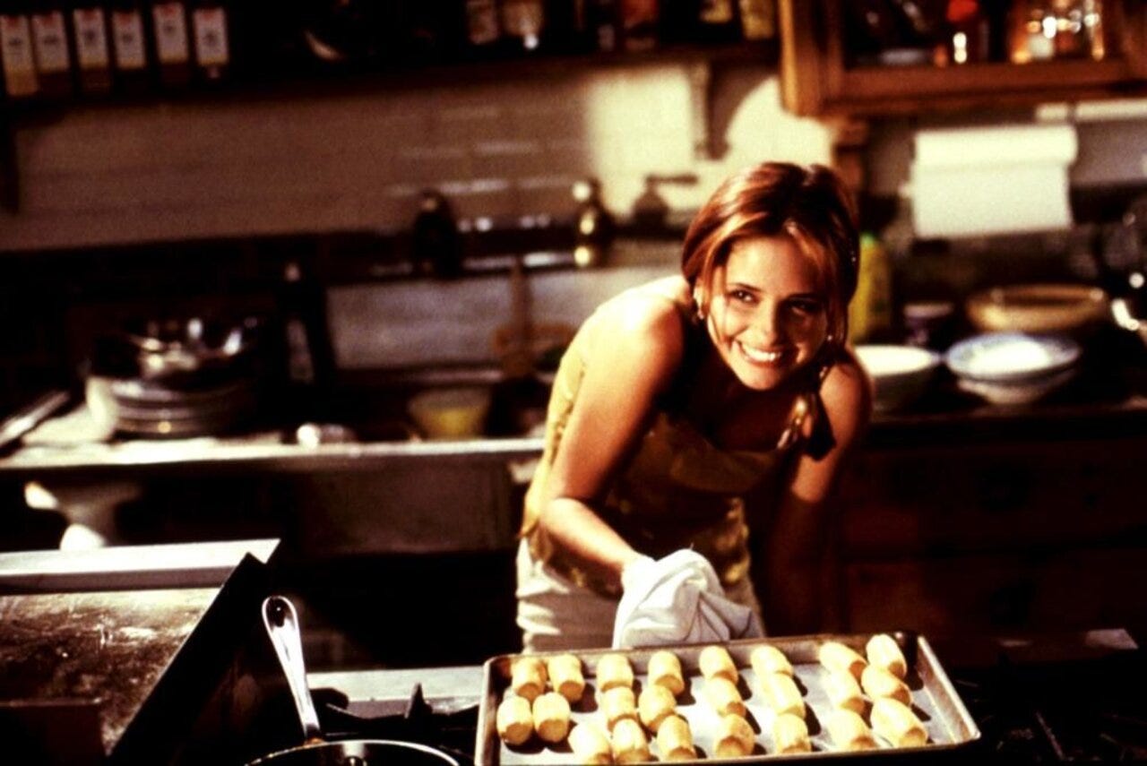Movie still from Simply Irresistible. A woman smiles in a restaurant kitchen in front of a tray of caramel eclairs.