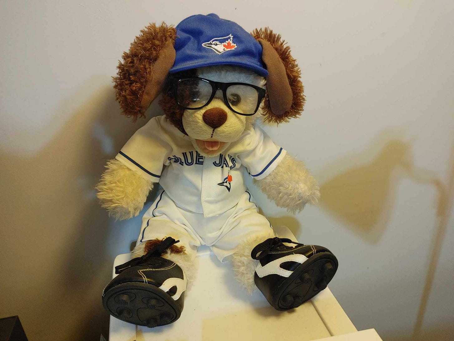 A build-a-bear dog wearing a Blue Jays had with its 2 brown floppy ears sticking out, along with a white Blue Jays jersey and pants over its white fluffy fur, and cleats on its feet.