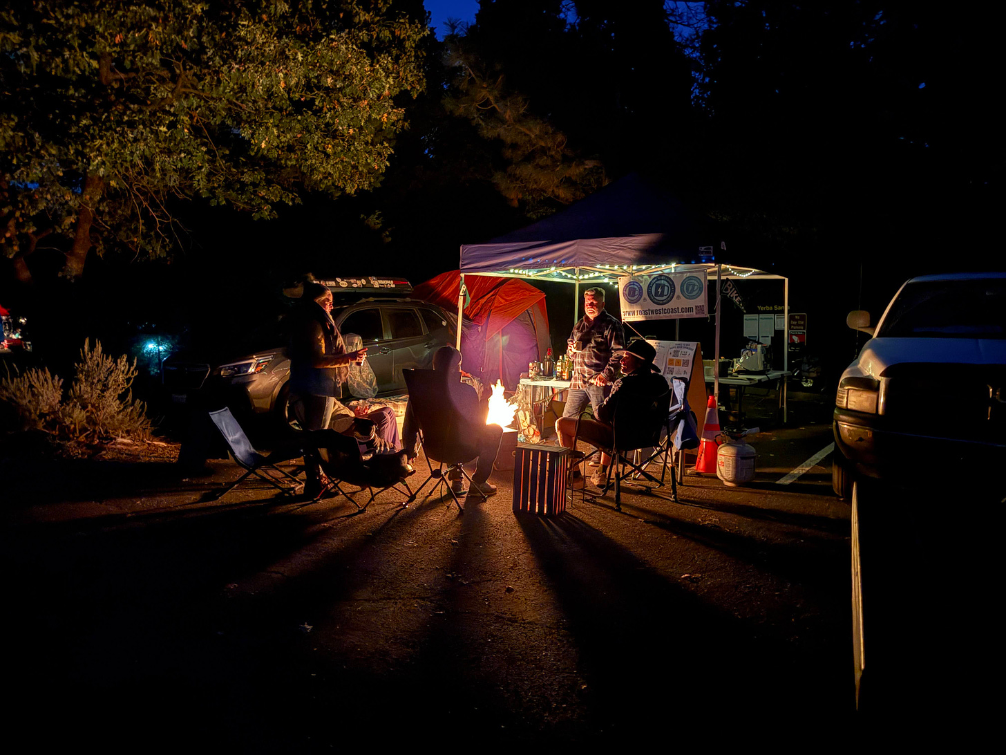 A nighttime photo of campers sitting around a blazing campfire in front of a pop-up tent. Trees are in shadow around the edges of the frame.