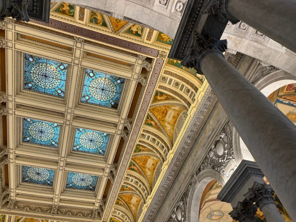 An ornate ceiling includes yellow frescos, blue stained glass squares, and marble crown moudling and arches and columns. 