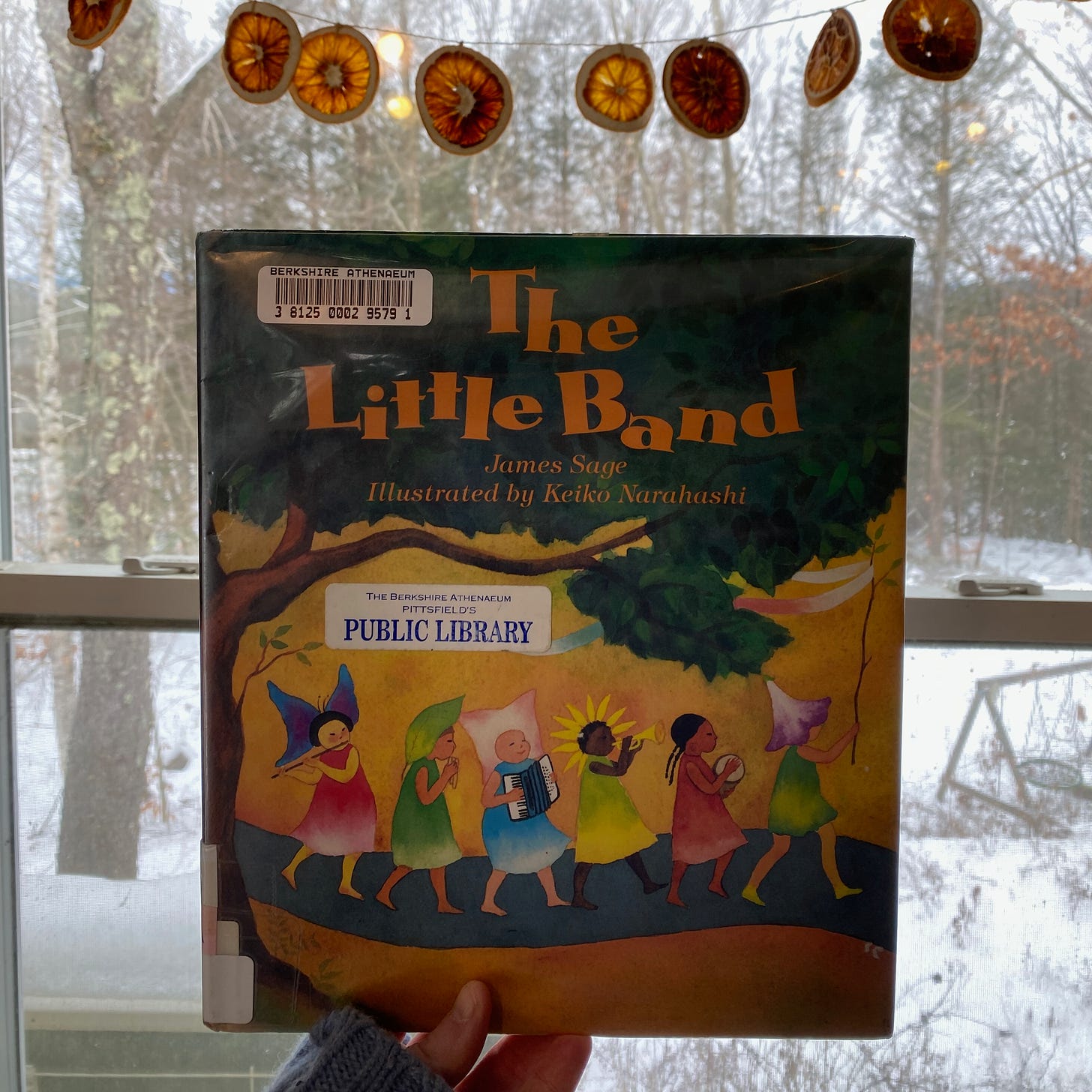 I’m holding The Little Band in front of a window underneath a garland of dried oranges.