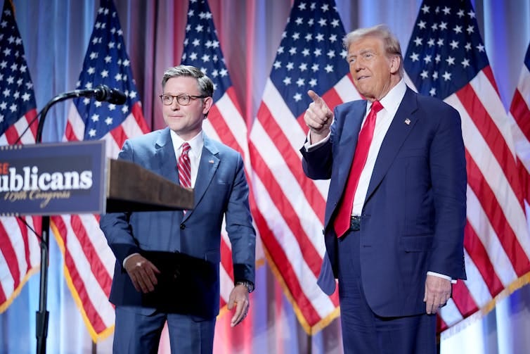 Two men in blue suits standing in front of many US flags.