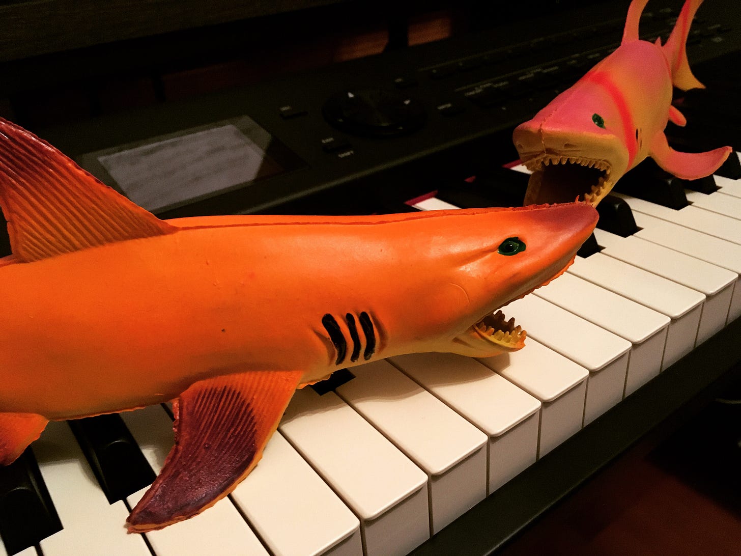Two orange and pink/purple toy sharks sitting atop a piano keyboard.