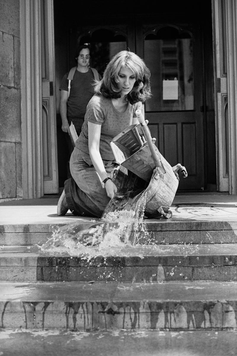 A black and white photograph of Mierle Laderman Ukeles in 1973. She is kneeling on the front steps of the Wadsworth Atheneum, pouring out the water from an old-fashioned metal mop bucket. The dirty wash-water is cascading down the steps.