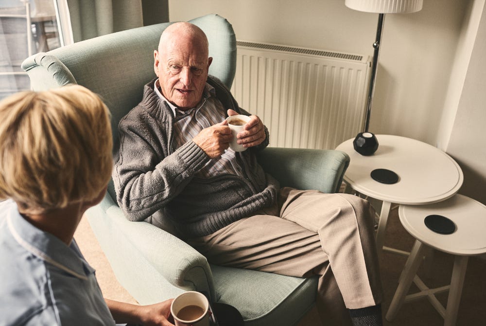 Female carer talking with older man and both having a cup of tea in his home