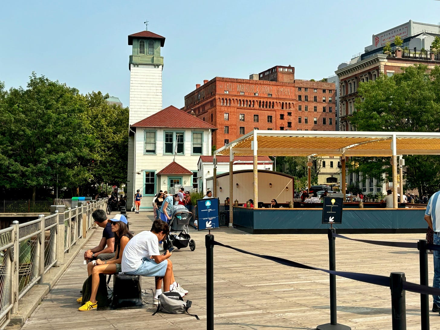 The line for the ferry. Facing Brooklyn, there is an ice cream shop in an old fashioned white building. Behind it is a brown building with lettering that reads THE EAGLE WAREHOUSE & STORAGE. The sun is shining and the trees are green.