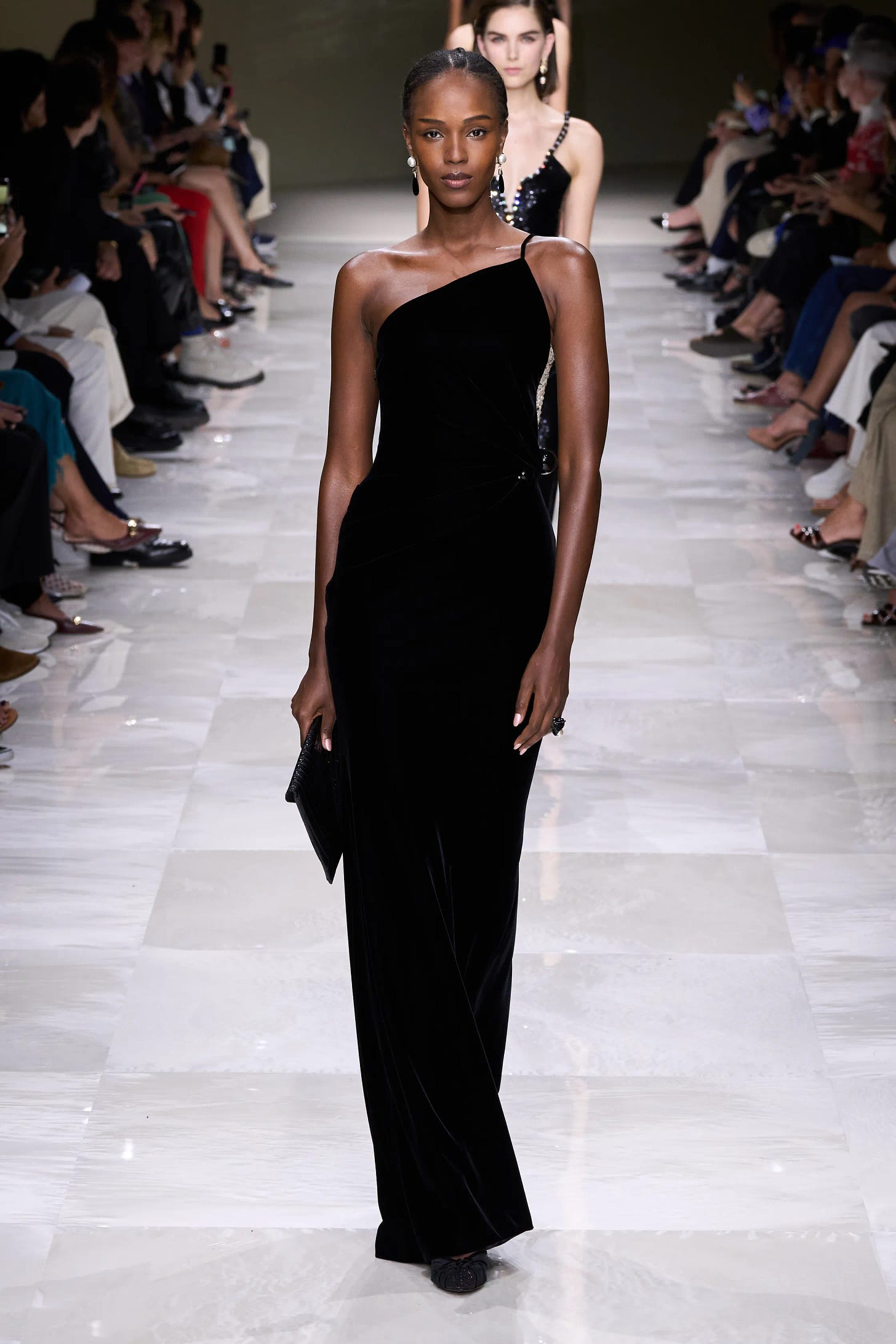 A dark-skinned model walks the runway wearing a slim one-shoulder gown in black velvet that fits through the waist and falls to the floor.