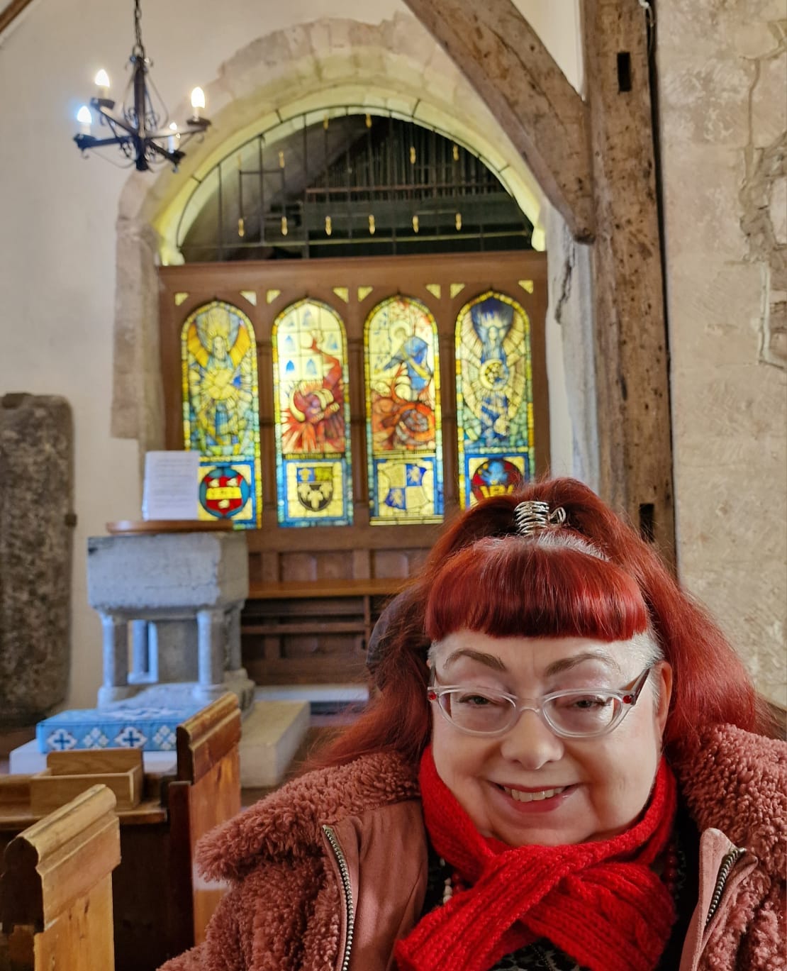 Penny is smiling, as she sits in her powerchair on a visit to a rural Norman-era church in West Sussex. The walls are mostly white and plain, with occasional stone arches preserved from Saxon times, and an ancient construction of wood beams that rise to the ceiling. In the background on the left, there is a white font. There is also a glimpse of the ‘slayers stone’ at the far left – this is connected to the folk tale of the water dragon, the knucker. Behind Penny there are four panels of modern stained glass windows, detailing the story of the knucker, which is mentioned in Penny’s post. The windows are brightly coloured with the knucker, a red snake-cum-dragon creature crossing the centre two panels with Jim Puttock offering him a poisoned pie. To the far left the glass panel represents the Angel of the Sun and on the far right the Angel of the Moon. Throughout there are wooden pews and other familiar churchy details. Penny has red hair in a ponytail, she wears glasses and a fluffy pink coat and small red scarf.