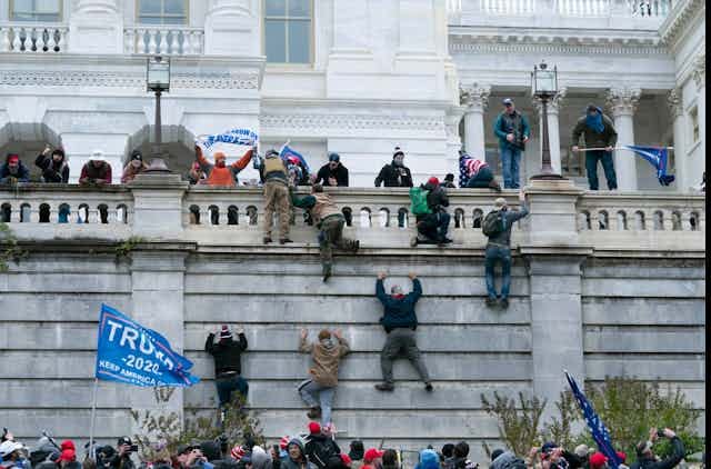 Men in military gear scale a wall with Trump 2020 flags flying in the foreground