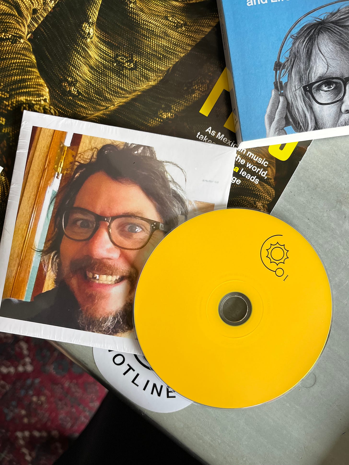 A Starship Casual CD, a yellow disc sitting on top of its paper sleeve, on a desk with a book and magazine nearby. The CD sleeve features a photo of Jeff smiling with a fake gold tooth and glasses.