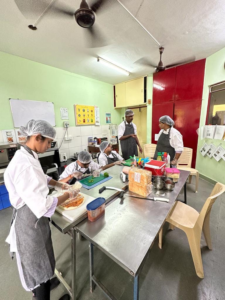 The image shows students in a bakery training unit, actively engaged in learning and practicing their skills. Each student is wearing a white chef's coat, apron, and hairnet. One student in the foreground is preparing a sandwich, while two others are discussing and assisting each other. In the background, a few more students are seated at a stainless steel table, chopping vegetables and preparing ingredients. The table is organized with various baking supplies, including bread and condiments. The room is well-lit, with instructional posters on the walls and a ceiling fan above. The atmosphere is collaborative and focused.