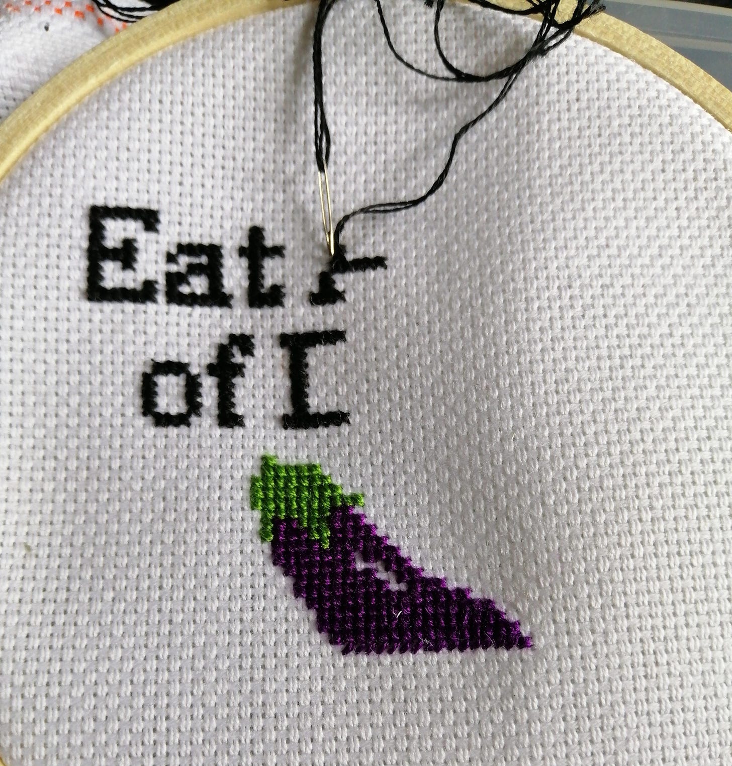 Cross stitch that is a work in progress. Text reads "eat a [blank] of D" and there is the top half of an eggplant below. 