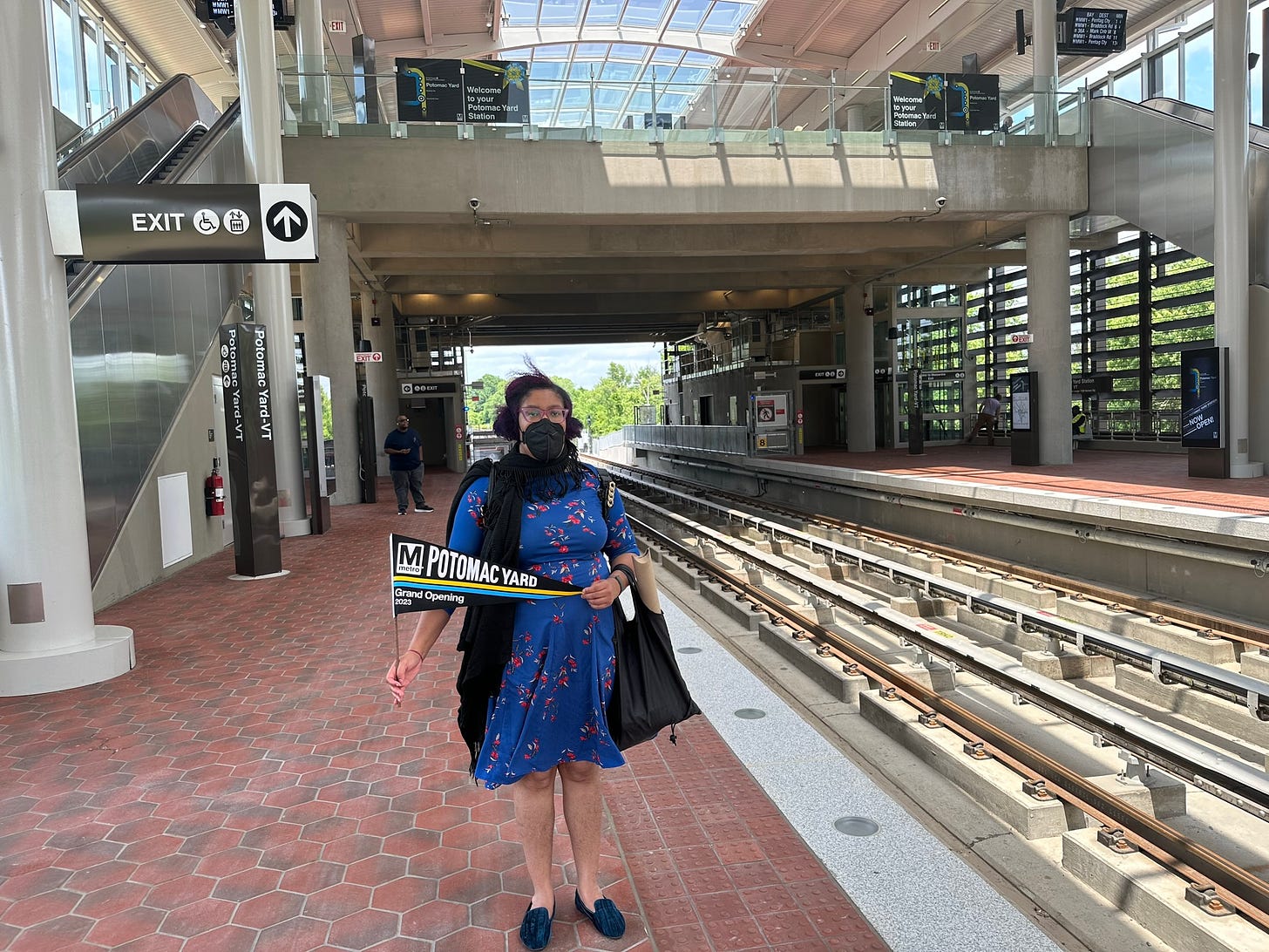Kristen is wearing a light-skin mid-size Black person wearing a blue floral dress and black N95 respirator  holding a black banner with Potomac Yard printed on it. She’s standing on a orange/grey hexagon tile pattern next to the new Potomac yard Metro tain tracks facing the camera. The station mezzanine is above her head and has openings to the  outside.