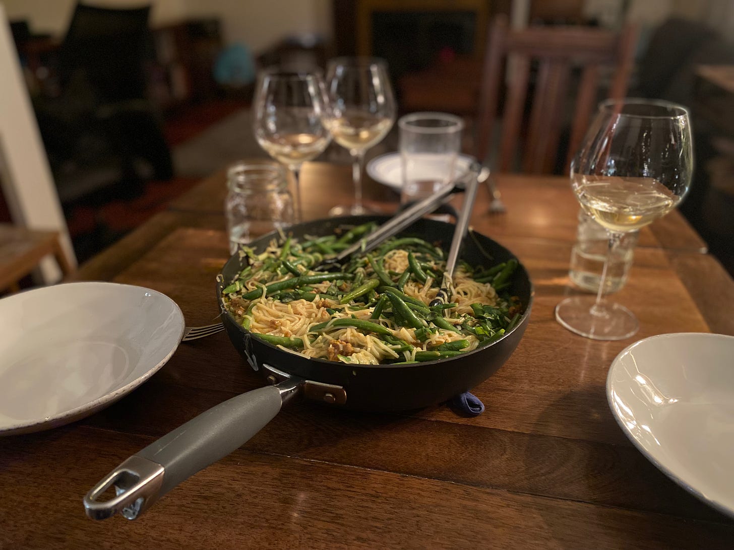 A table set with white plates and wine glasses, partially full with white wine. A sauté pan of the noodle dish described above sits on a potholder in the centre, a pair of tongs sticking out of it.