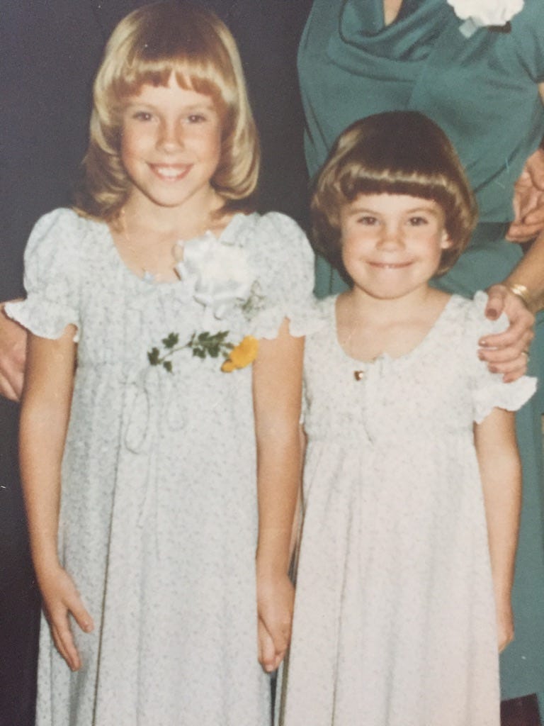 Two young, smiling sisters. One is blonde and taller. The other is brunette and shorter. They're wearing matching dresses.