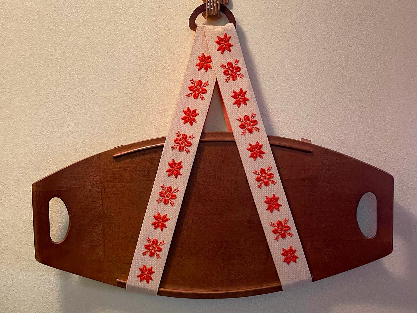 A wooden tray hangs from two strips of ecru fabric embroidered with orange snowflake-like designs. 