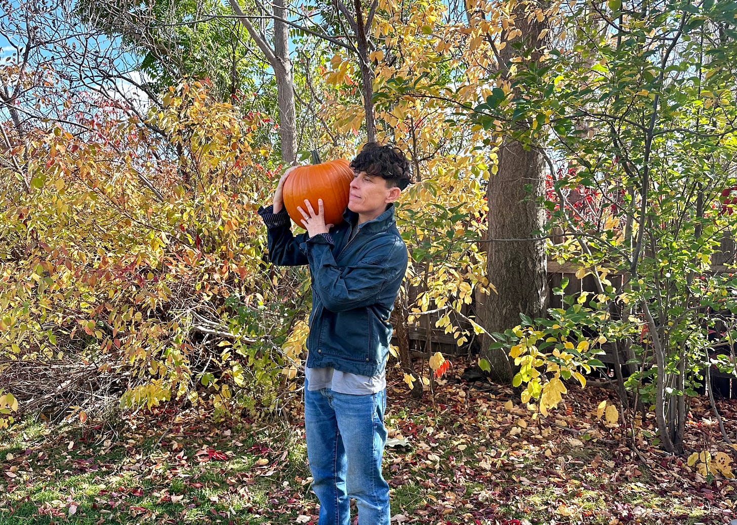 Andrea Gibson standing in front of fall colored trees with yellow, red and green leaves, holding a giant pumpkin on their right shoulder and smiling. Andrea is wearing a blue worker jacket and blue jeans and smiling. 