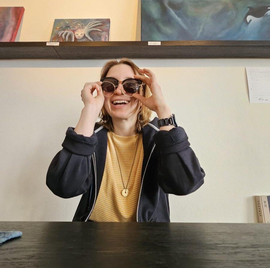 A petite white woman with straight, shoulder length brown hair, wearing a navy hoodie over a yellow shirt. Maria is sitting in a cafe wearing sunglasses and glasses at the same time, looking at and talking to the person in front of her.