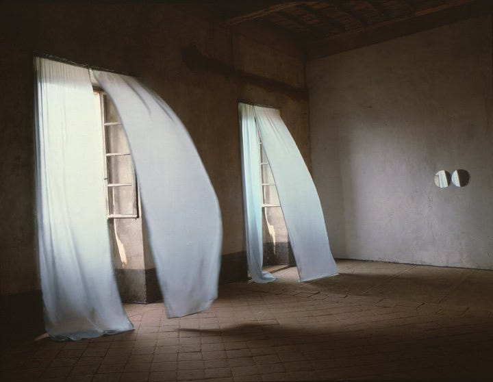 Felix Gonzalez-Torres’s work "Untitled" (Loverboy), 1989. as installed in Itinerari. Castello di Rivara, Turin, Italy. There are two open windows bringing light into an otherwise dim room. Pairs of pale blue curtains are lifted by the breeze coming in through the windows.