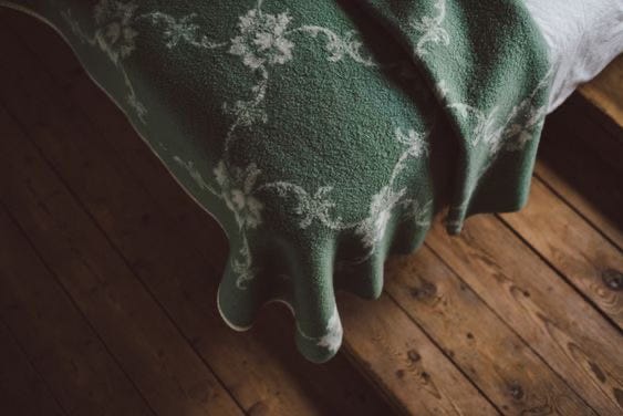 A green blanket with white floral patterns drapes over the edge of a bed, contrasting with the rustic wooden floor beneath.