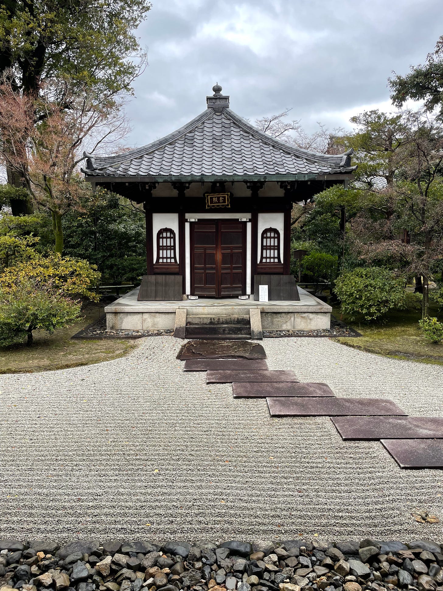 A small Japanese temple building sits behind a zen rock garden surrounded by bushes and trees