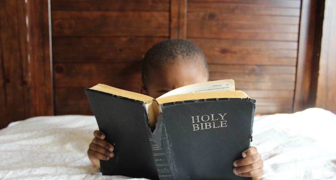 boy reading Holy Bible while lying on bed