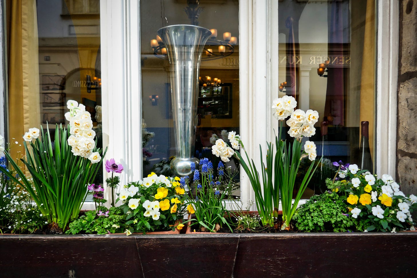 A window in front of a small shop in Germany with a window box full of blooming flowers. You can see a tall glass vase in the window and furnishings in the room.