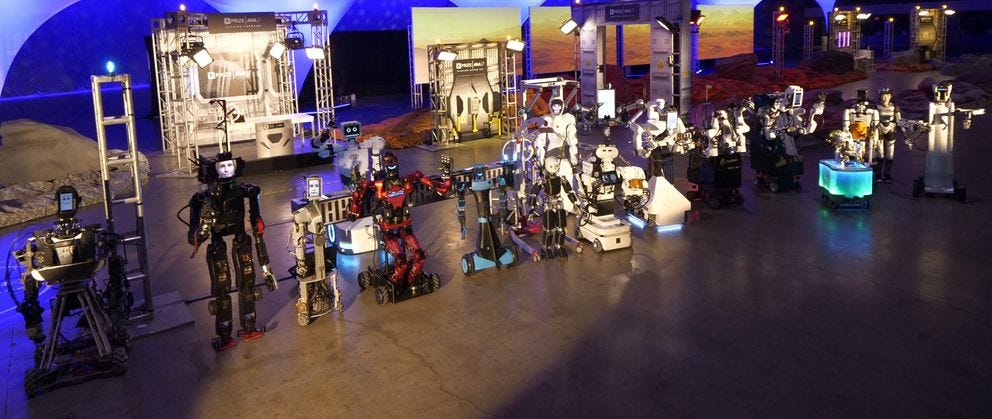 A wide angle photo showing a dozen different robots lined up next to each other in a competition arena.