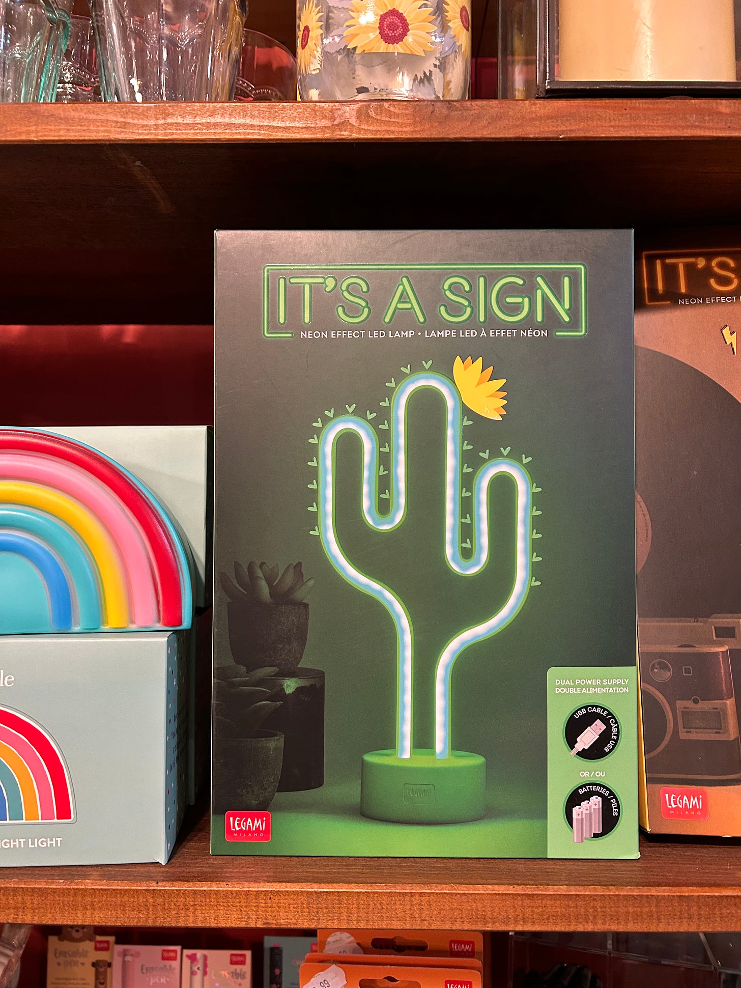 A box sitting on a shelf that shows a bright green neon lamp in the shape of a cactus. Above the neon lamp are the words IT'S A SIGN in bright green block lettering.