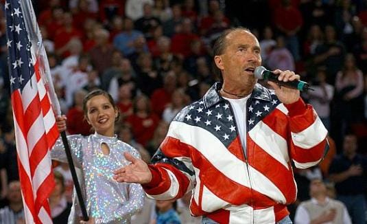 Photo of Lee Greenwood dressed in an American flag jack, holding a microphone and singing in front of a crowd