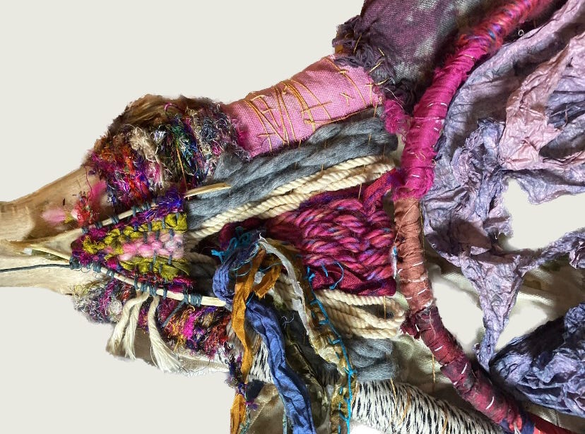 A colorful textile art piece made from various fabrics, yarns, and threads. The piece features a central wooden bone-like structure wrapped with different textured and colored materials. Fabrics in shades of purple, pink, and blue are prominently visible, along with various knots, weaves, and embellishments.