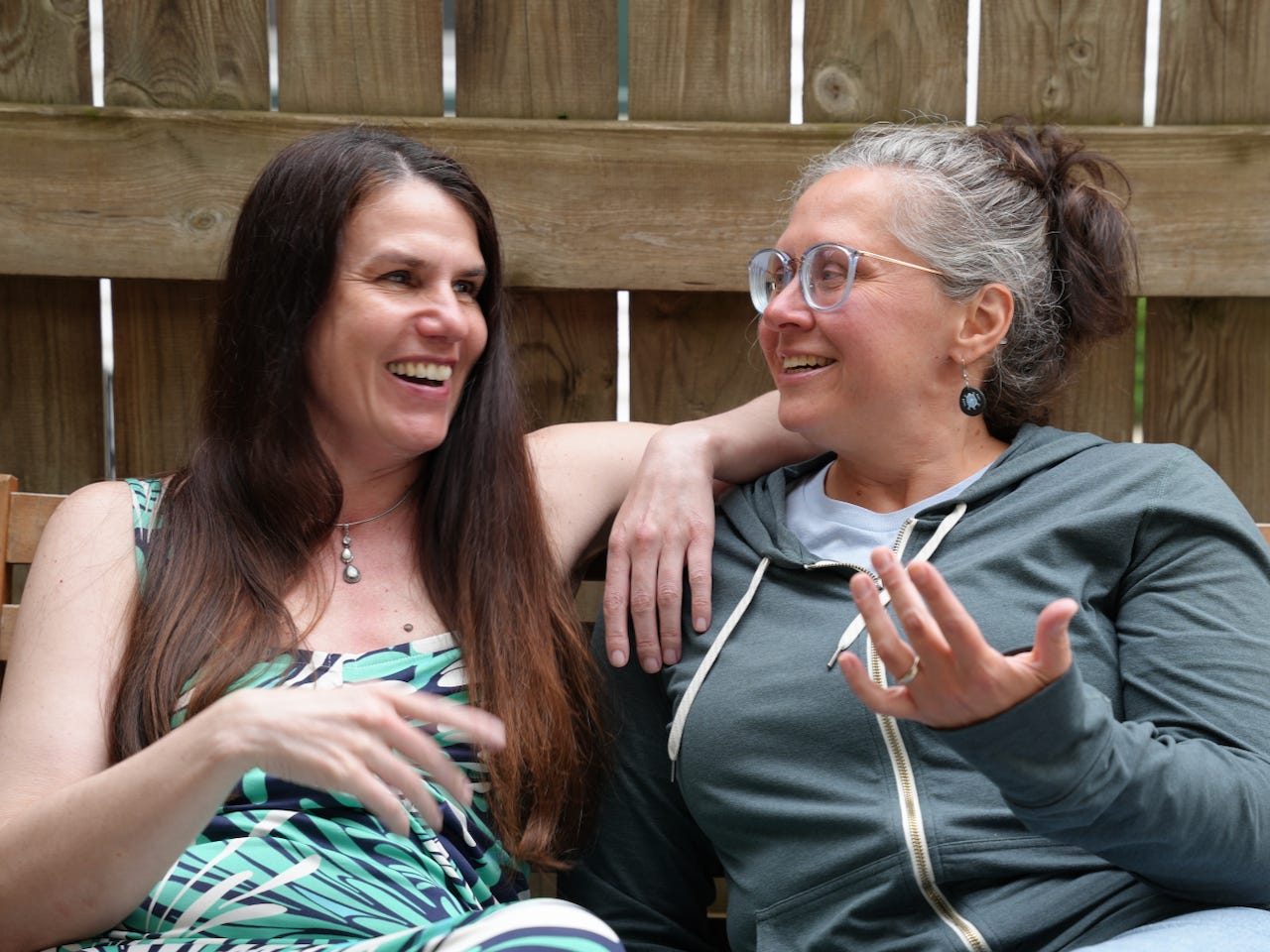 Two women (Alexandra Hidalgo and Kristin Arola) sit together and smile as they talk. Their arms are across each other. There’s a fence behind them. It is daytime. Two women (Alexandra Hidalgo and Kristin Arola) sit together and smile as they talk. Their arms are across each other. There’s a fence behind them. It is daytime.