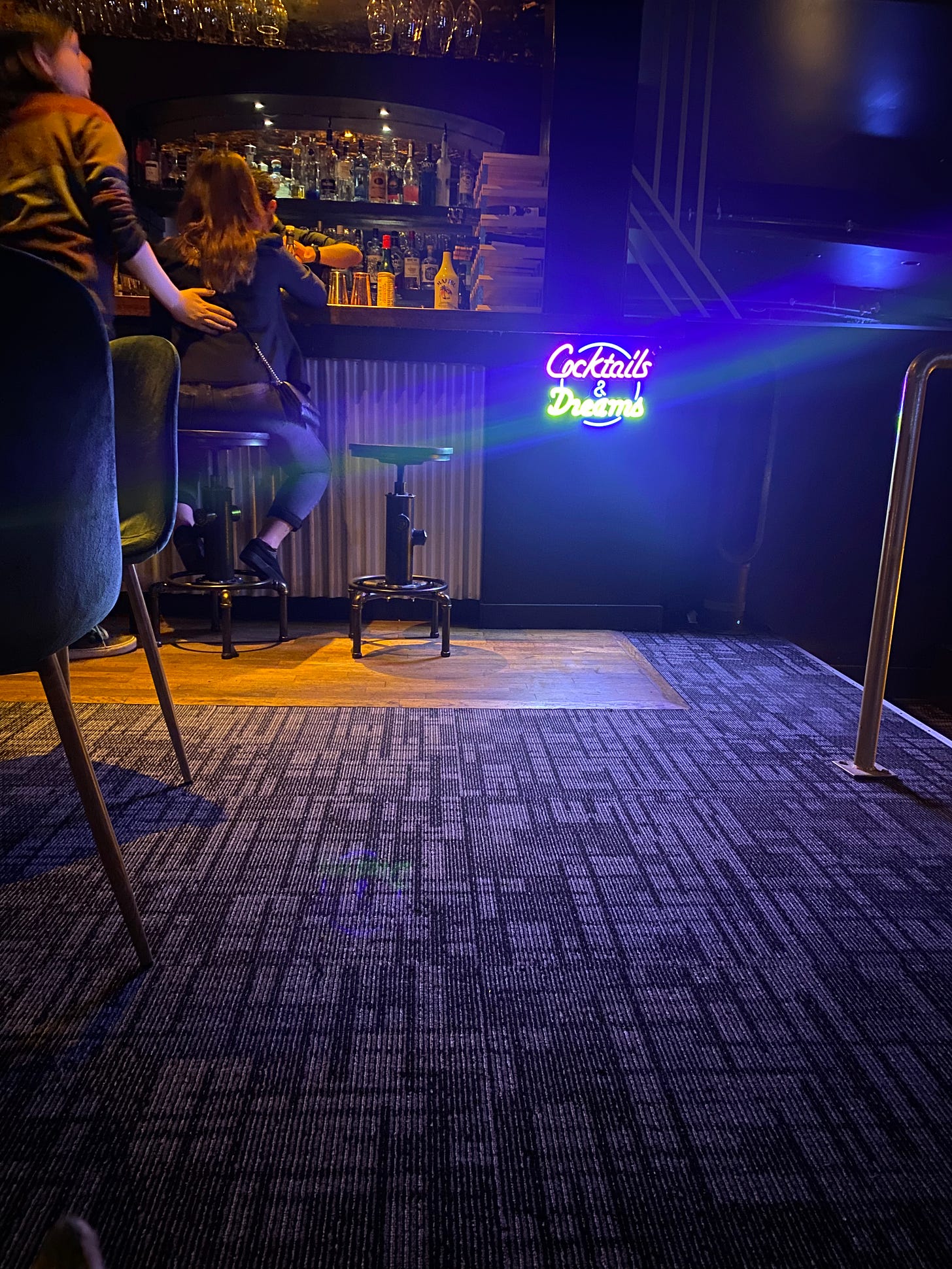 A barroom bathed in purple-blue light. The focus is on a neon sign on the bar front that reads 'cocktails & dreams' in pink and green. Next to it, a person standing has his hand on the back of a person sitting at the bar. In the foreground are blue velvet chairs and a geometrically patterned carpet.